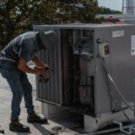 A worker in a bucket hat repairs an outdoor air conditioning unit on a rooftop. - Photo by José Andrés Pacheco Cortes on Pexels