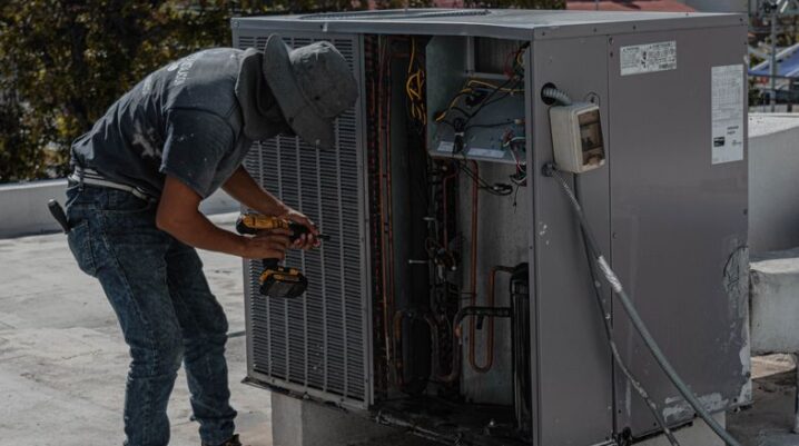 A worker in a bucket hat repairs an outdoor air conditioning unit on a rooftop. - Photo by José Andrés Pacheco Cortes on Pexels